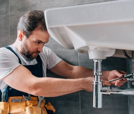 A professional plumber repairing a leaking basin pipe under a bathroom sink.