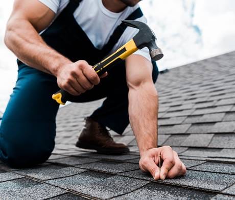 A man repairing the roof of a house, using tools and safety gear under bright daylight.